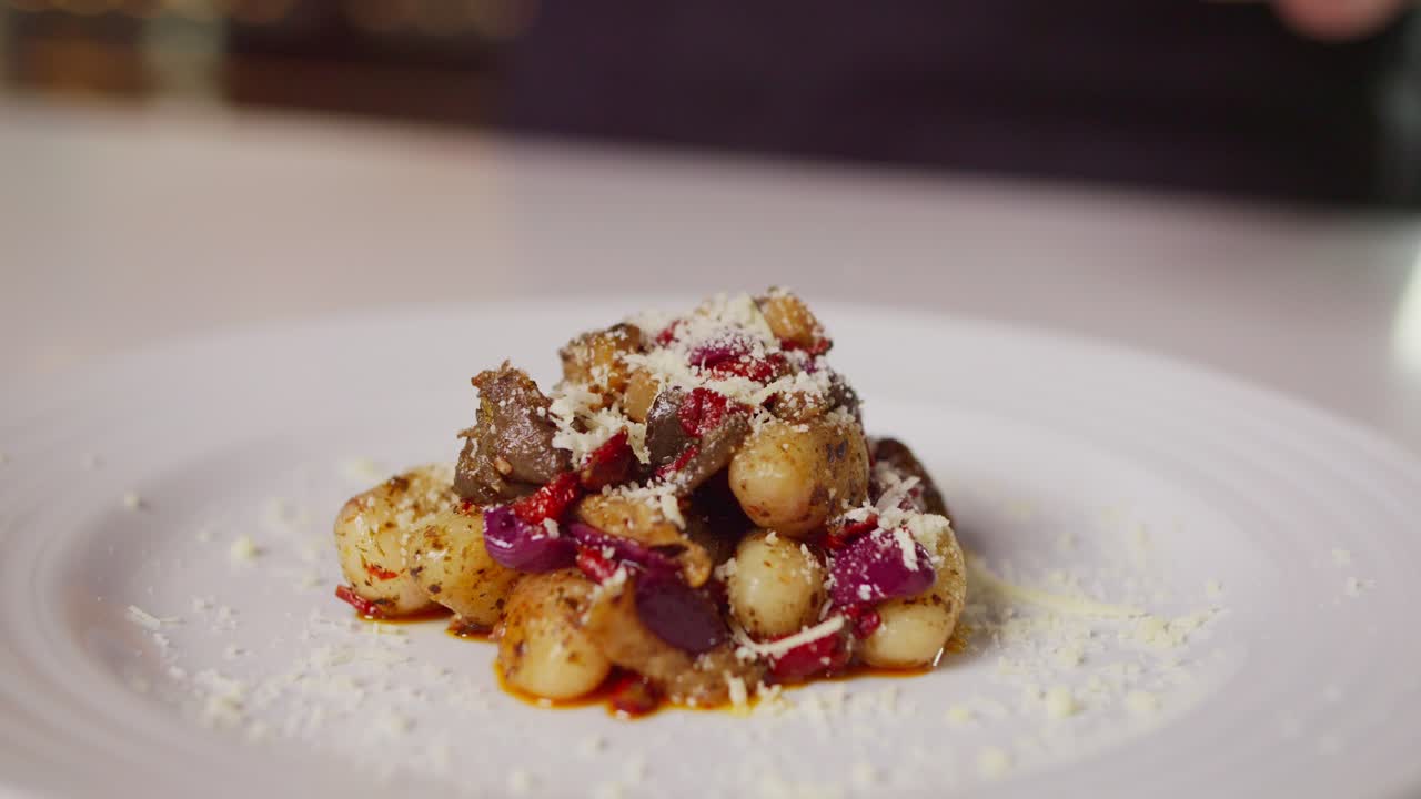 A close-up of gnocchi topped with meat, red onion, and grated cheese on a white plate, with cheese being freshly sprinkled from above, creating an elegant and appetizing presentation.