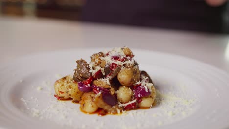 a close-up of gnocchi topped with meat, red onion, and grated cheese on a white plate, with cheese being freshly sprinkled from above, creating an elegant and appetizing presentation.