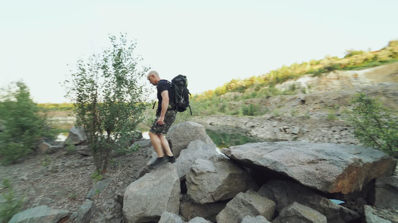 adult traveler with backpack is examining the terrain by the river and walking along the rocks along the bank on the background of a wonderful landscape