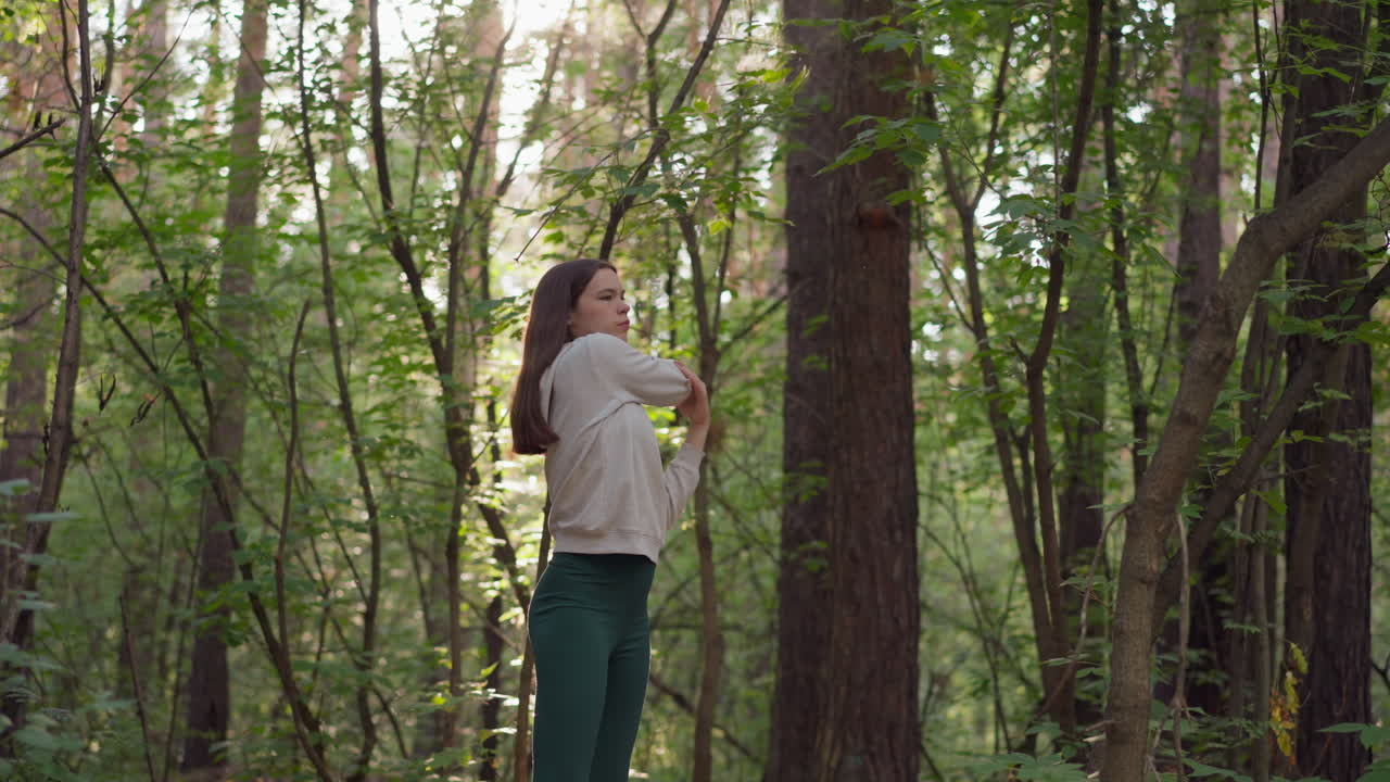 젊은 여성 운동 선수가 숲에서 팔을 펴고, 다양한 종류의 나무로 둘러싸인 스포츠를 하고, 은 날씨에 야생에서 태양 광선이 여인에게 떨어집니다.