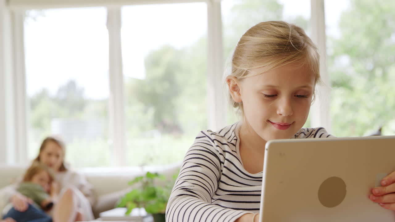 niña en la mesa con tableta digital escolarizando en casa durante la pandemia de salud con su familia en el fondo
