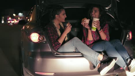 Handsome-girlfriends-sitting-in-the-open-trunk-of-the-car,-talking-and-taking-sandwiches,-drinking-from-the-paper-cup-during-the