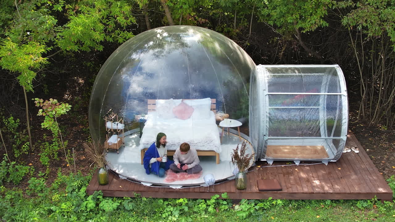 Gennemsigtigt bobletelt ved glamping, frodig skov rundt om og indvendigt. Et par sidder under et tæppe, holder kopper og snakker