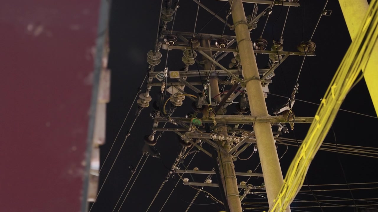 Night utility pole with tangled cables, lamp glow, transformer hardware, intersecting wires, urban skyline edge, quiet moody atmosphere, industrial texture, maintenance perspective, cinematic