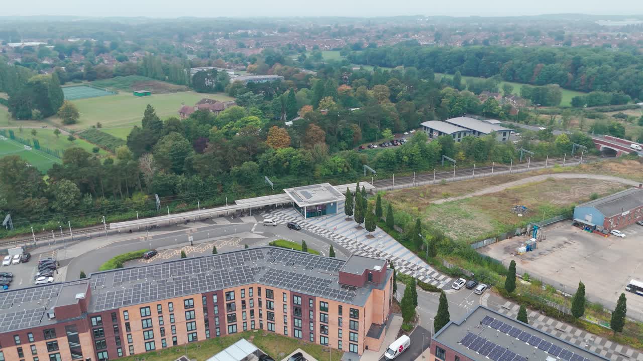 Aerial view of an urban landscape featuring buildings, trees, cars, and a parking lot. The area includes roads, railway tracks, and greenery, highlighting infrastructure
