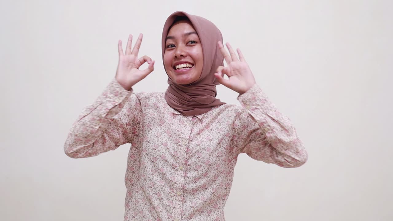 Wanita muda muslim Asia yang bersemangat berdiri menunjuk ke bawah dan menunjukkan isyarat tangan 'oke'.