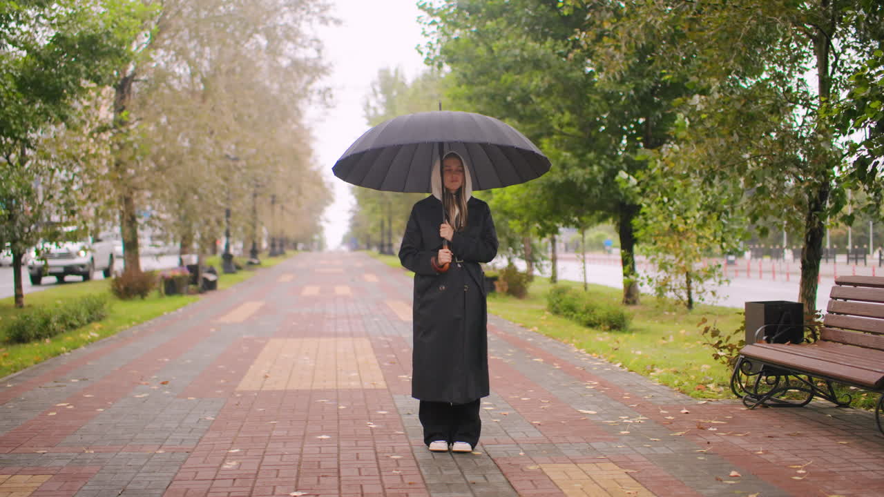 Woman in black trench coat holding umbrella standing in middle of empty city walkway on rainy autumn day, trees and benches along path, urban solitude atmosphere with calm mood and overcast weather