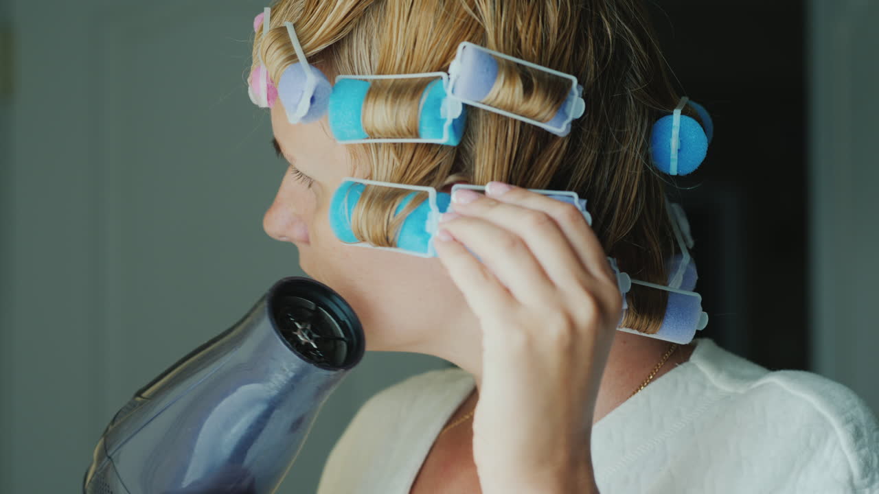 한 젊은 여성이 헤어스타일에 모양을 부여하기 위해 머리에 헤어드라이어 컬러로 머리를 말립니다.