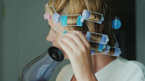 a young woman dries hair with a hairdryer curlers on her head to give shape to her hairstyle