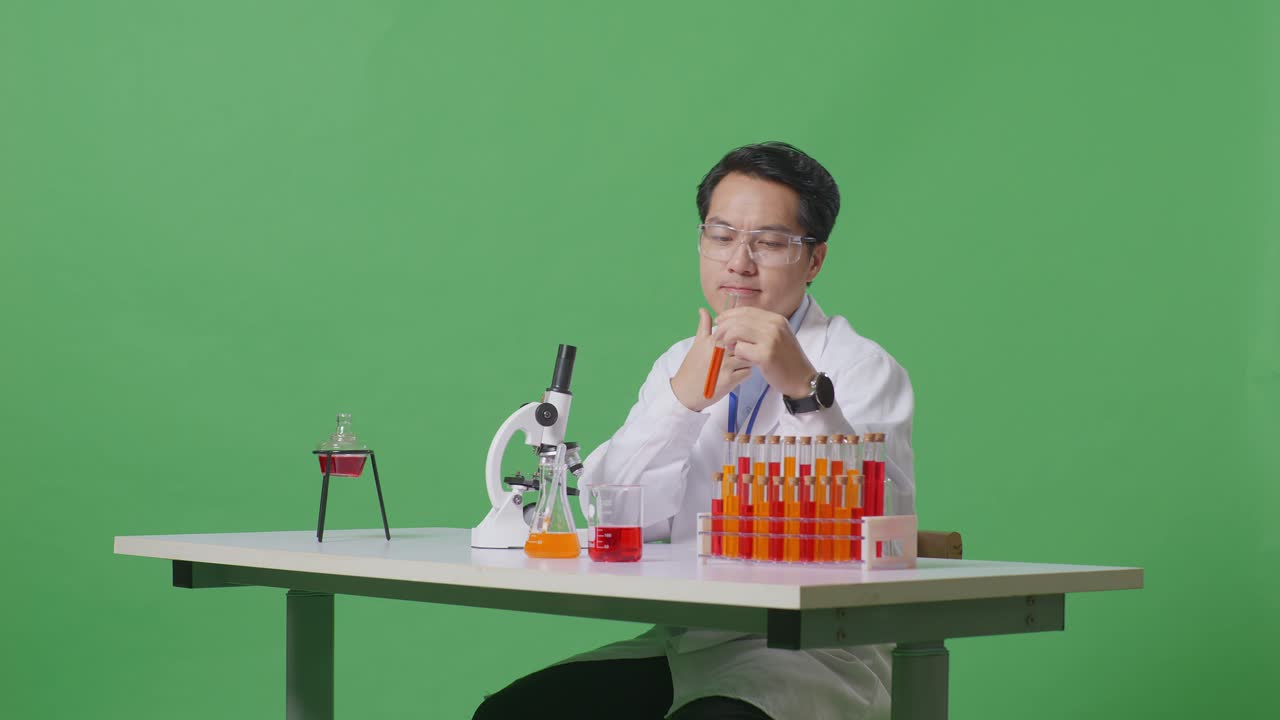 아시아 남성 과학자가 실험관에서 생각하고, 주위를 둘러보고, 지표손가락을 올리며 녹색 스크린 백그라운드 실험실에서 현미경으로 테이블에서 작업합니다.