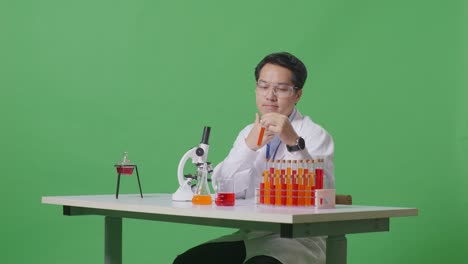 아시아 남성 과학자가 실험관에서 생각하고, 주위를 둘러보고, 지표손가락을 올리며 녹색 스크린 백그라운드 실험실에서 현미경으로 테이블에서 작업합니다.