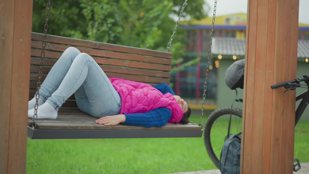 Mulher sentada ao ar livre tranquilamente, mulher serena descansando em um balanço, mulher calma relaxando em um balanço de parque com uma bicicleta por perto, mulher tranquila de jaqueta rosa aproveitando um momento de tranquilidade em um balanço de parque