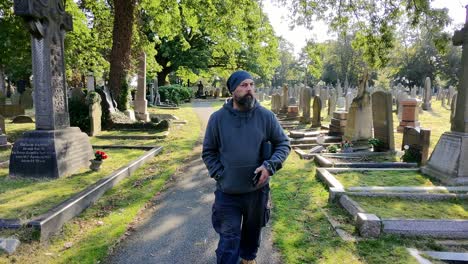 bearded male wearing hat and casual clothing walking alone in peaceful sunlit cemetery