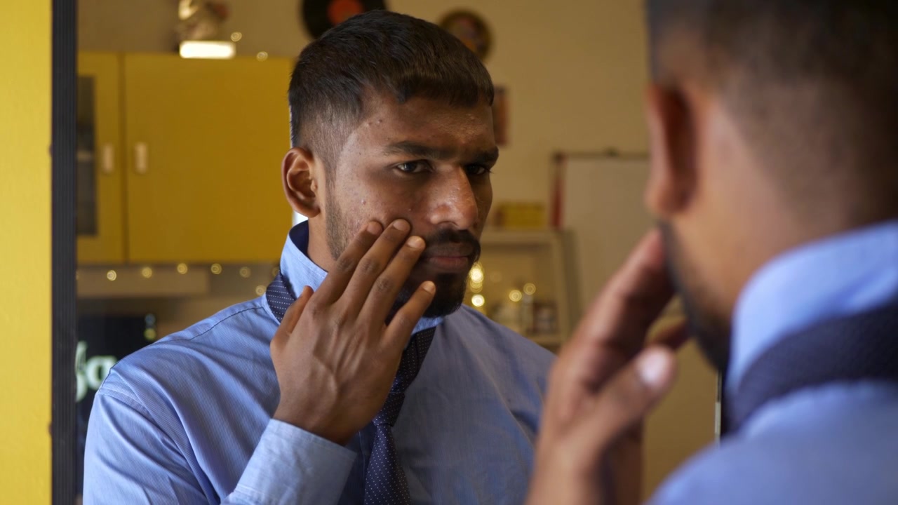 Giovane uomo indiano in abbigliamento da ufficio/universitario si sistema la cravatta mentre si guarda allo specchio, insoddisfatto della pelle abbronzata e piena di acne; un momento intimo in camera da letto che riflette preoccupazioni sull'immagine di sé e insicurezza