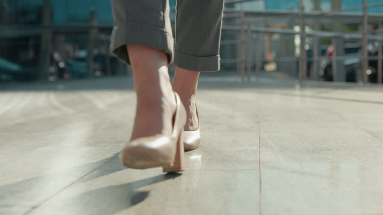 Close up leg view of confident lady walking gracefully in nude high heels on sunlit walkway with modern building, iron rail, parked cars in blurred background