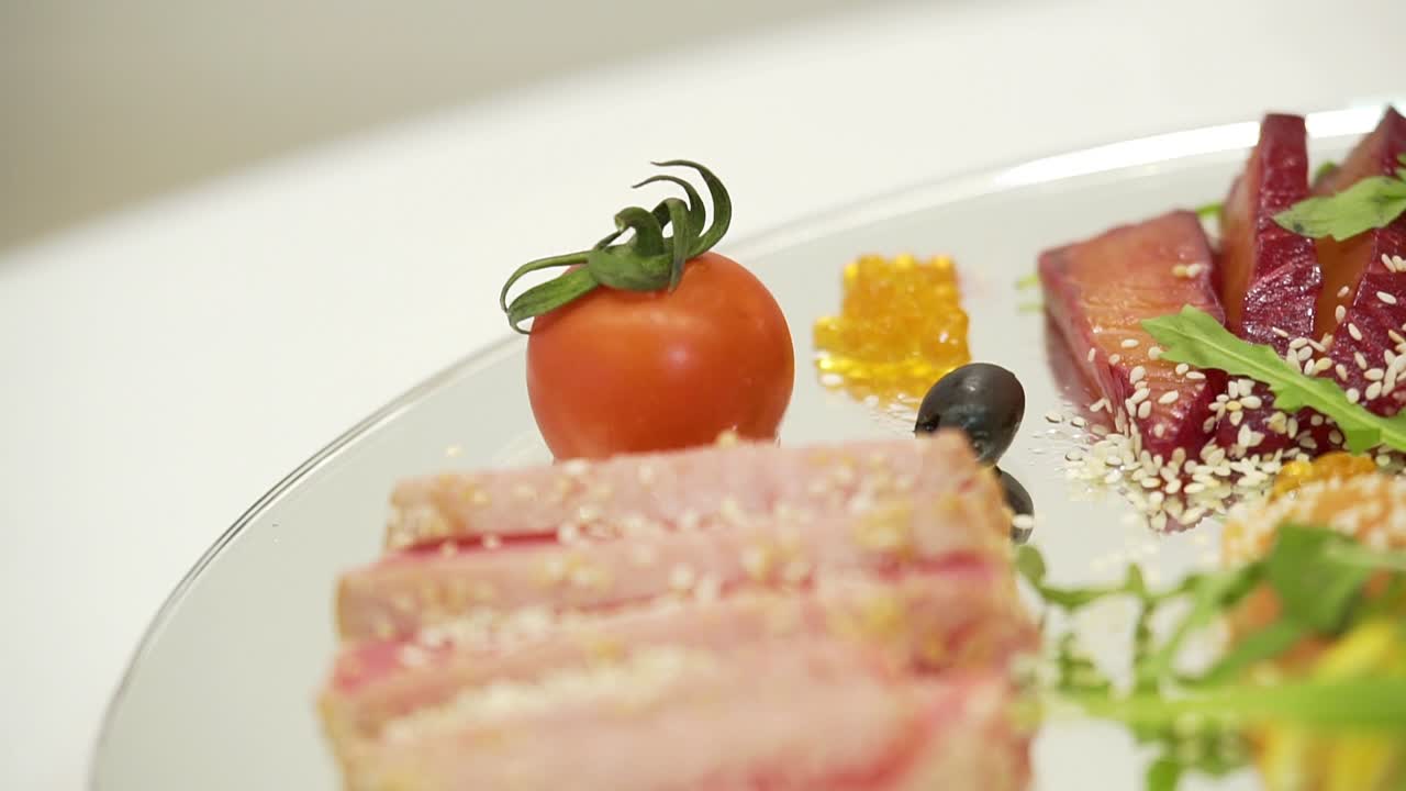 Sliced bacon with sesame is decorated with cherry tomato near pieces of dry-cured meat and lemon with olives and cilantro on a transparent plate on the blurred background in the cafe.