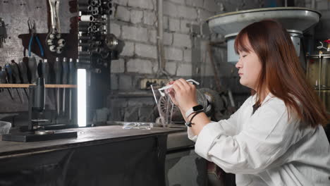 lab assistant in white coat picks up protective face shield, puts it on, and relaxes back on seat, with partial view of another person nearby, light, machinery, and tools in industrial workshop