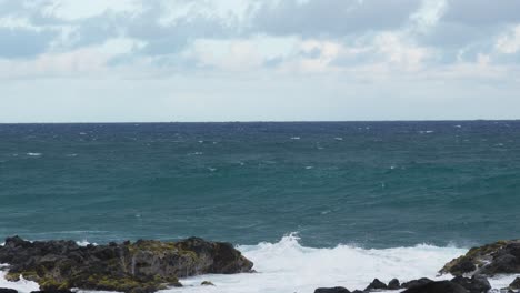 Wide ocean view with rolling waves breaking against jagged black rocks, foamy surf spreading along the rugged coastline under a bright but cloudy horizon