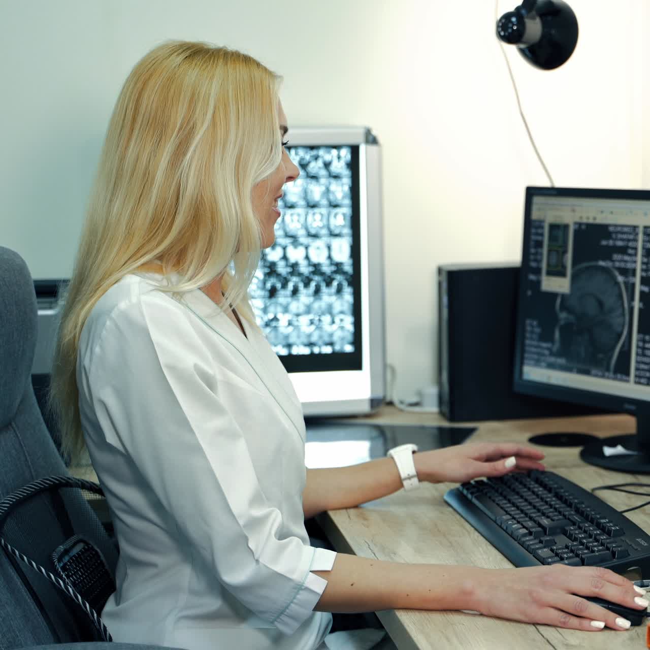 Blonde smiling female medic sitting at the table in front of computers. Lab technician looking through the head scan of a patient
