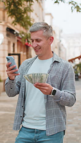 happy rich man counting holding money dollar cash using smartphone calculator app on city street