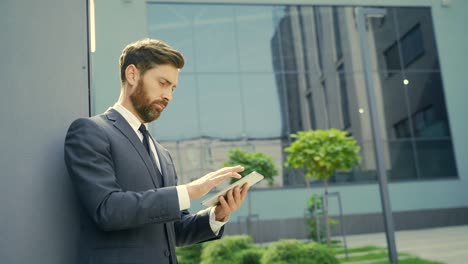 stylish bearded businessman in formal business suit standing working with tablet in hands on background modern office building outside.