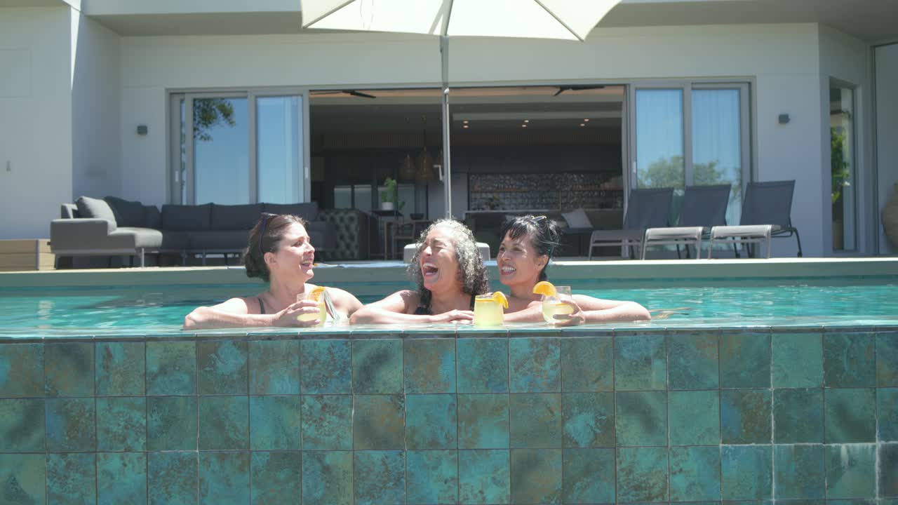 Diverse female friends in swimsuits relaxing chatting with drinks as camera closing in at pool