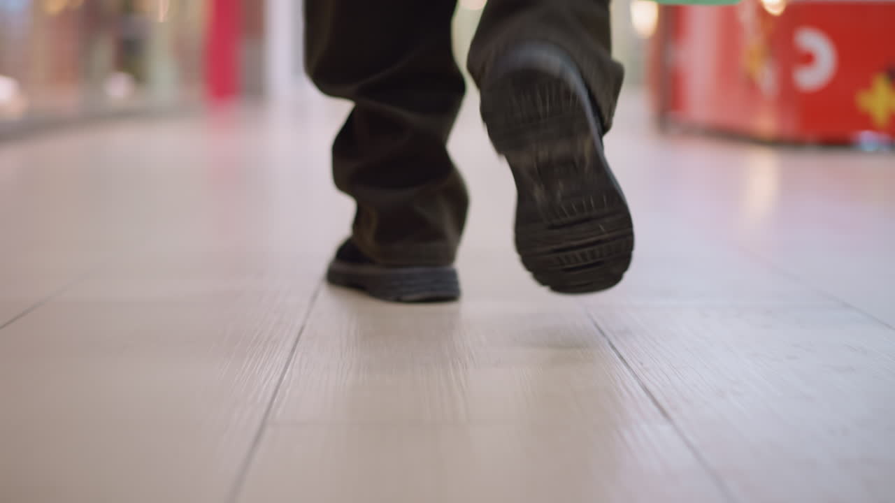 low angle shot showing legs and shoes of person walking through modern shopping mall carrying green bag focus on movement on polished tile floor blurred store displays background urban retail environment