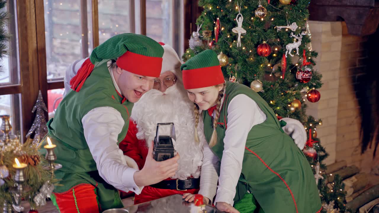 Santa with elves doing selfie. Joyful Santa together with fairy characters making photo on Christmas background indoors. New Year holidays.