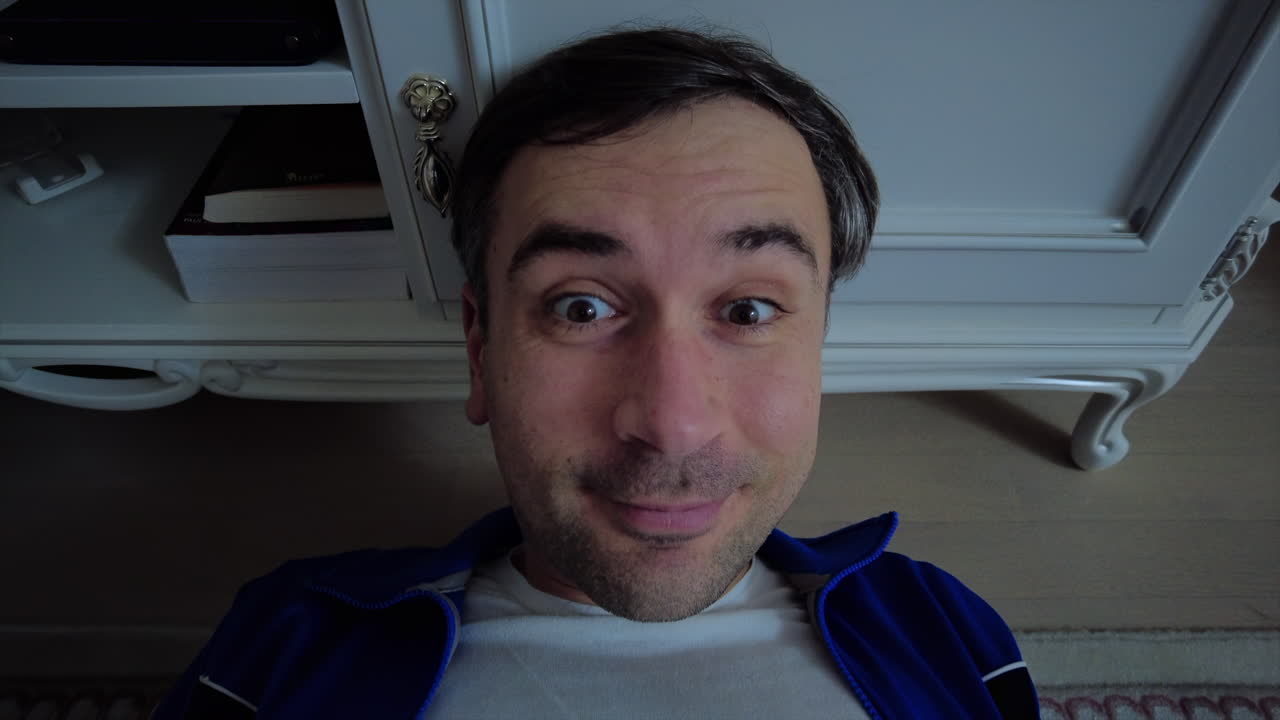 A man lies on the floor in a cozy room, smiling widely at the camera. His relaxed posture suggests he is enjoying a calm and joyful moment at home