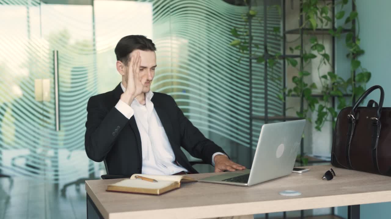 잘생긴, 젊은, 세련된 남자가 비즈니스 의상을 입고 멋진 사무실에서 책상에 앉아있는 동안 노트북으로 비디오 통화를 시작합니다.