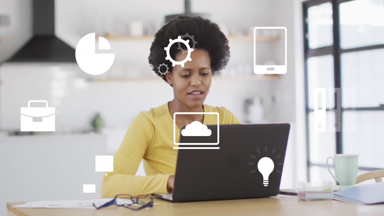 Animation of icons over african american woman using laptop in kitchen