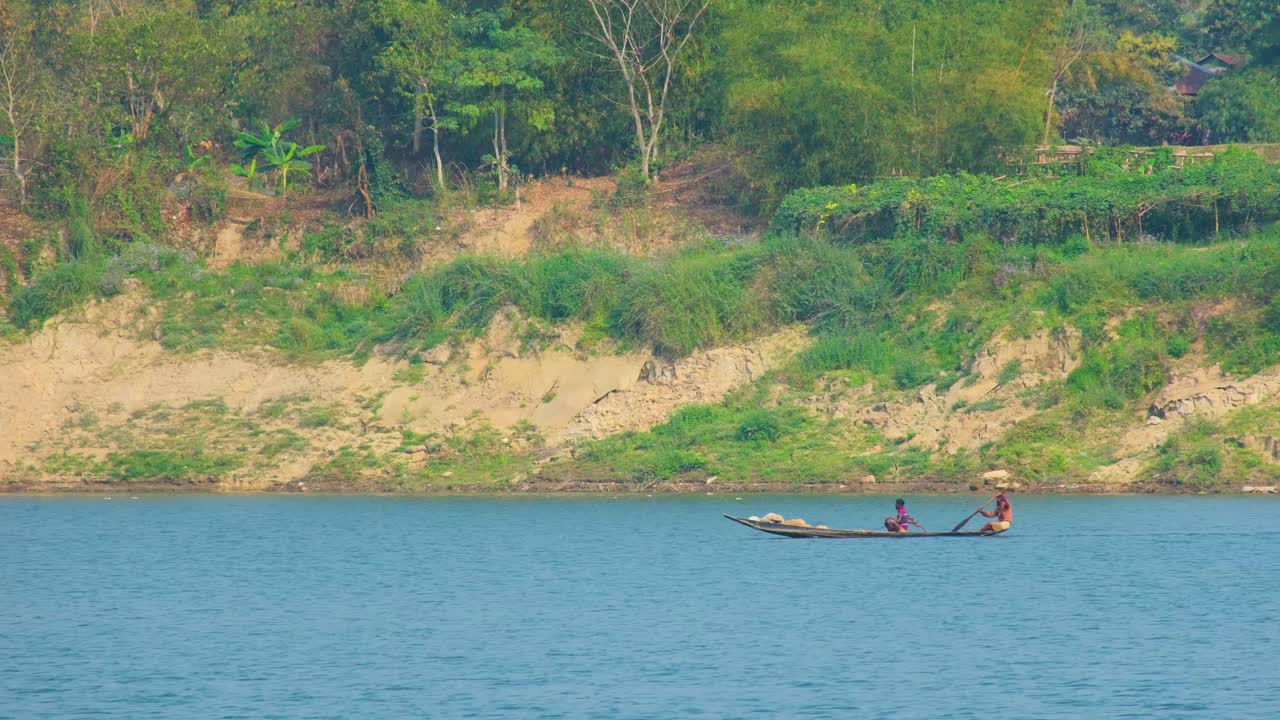 Dois homens asiáticos remam em um pequeno barco tradicional no rio, em plano médio.