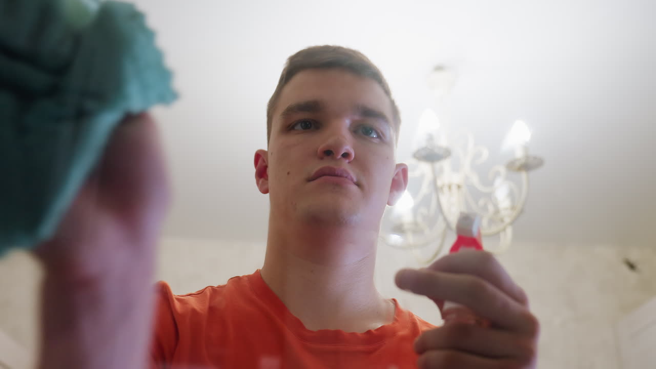 Young man in orange shirt holding spray bottle and applying cleaning solution onto bathroom mirror, focusing on household care, hygiene, removing dirt, streaks and water stains