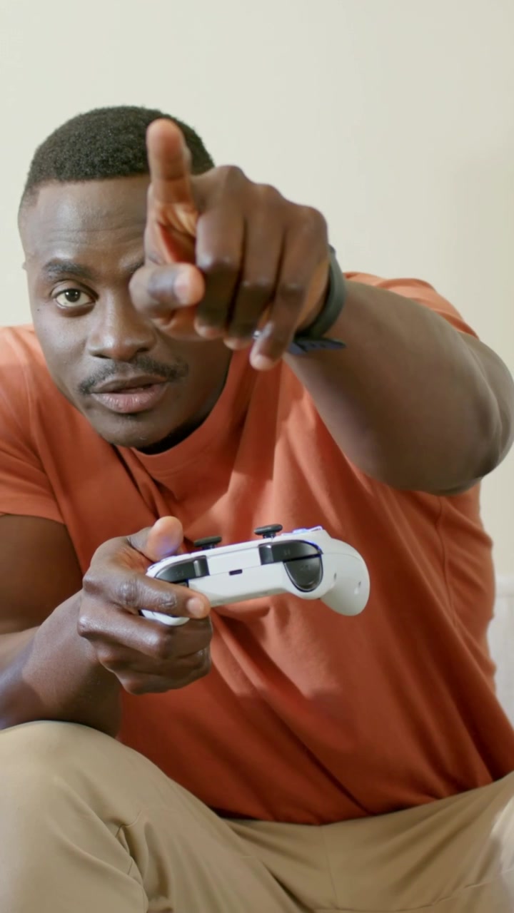 Vertical video: Reacting to game cue, gamer in rust tee pointing on couch, holding white controller