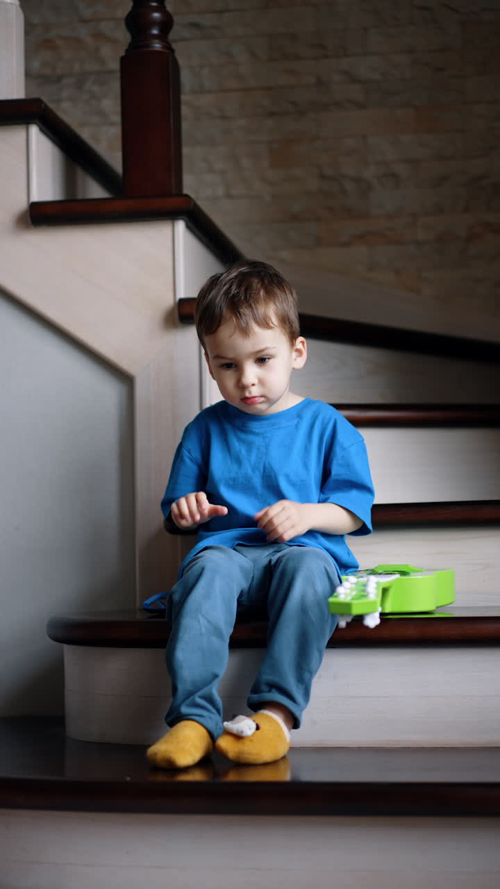 Calm Caucasian toddler sitting on the stairs at home. Baby boy puts off his sunglasses, stands up and walks away. Vertical video.