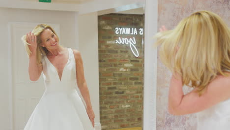 smiling mature bride looking at wedding dress in mirror of bridal store