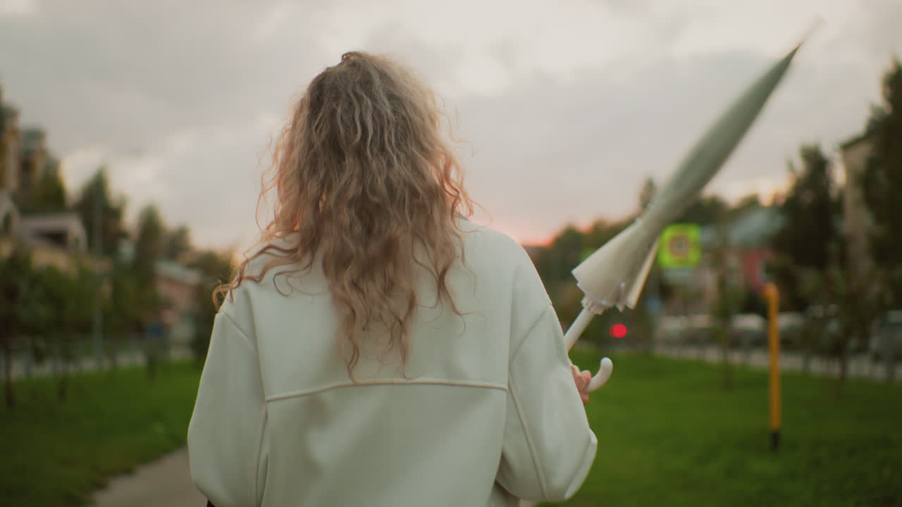 rear view of girl wearing white coat walking along paved path twirling umbrella playfully during soft sunset with blurred signpost, passing car with red light and urban environment