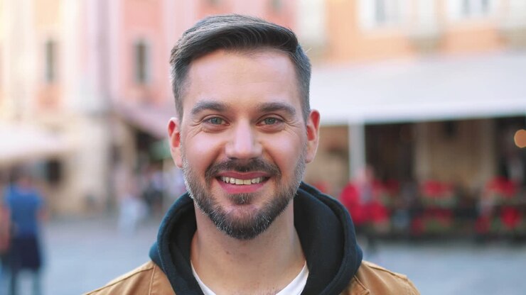 Primo piano di un uomo caucasico con la barba che guarda la telecamera e sorride per strada.
