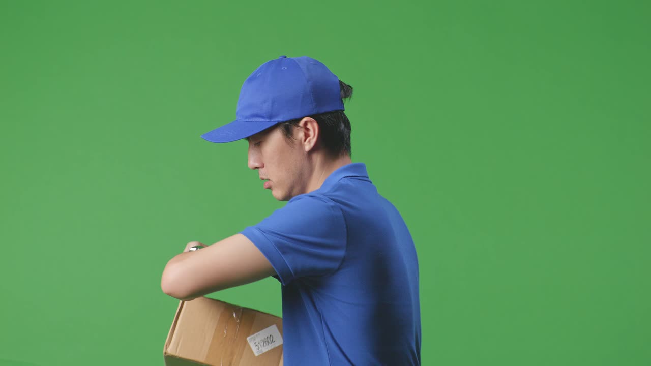 Close Up Side View Of Asian Male Courier In Blue Uniform Checking Watch And Running While Delivering A Carton On Green Screen Background In The Studio