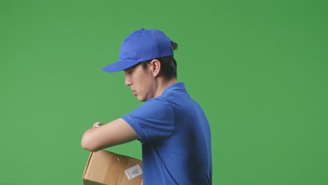 vista lateral de cerca del mensajero masculino asiático en uniforme azul comprobando el reloj y corriendo mientras entrega una caja de cartón en el fondo de la pantalla verde en el estudio