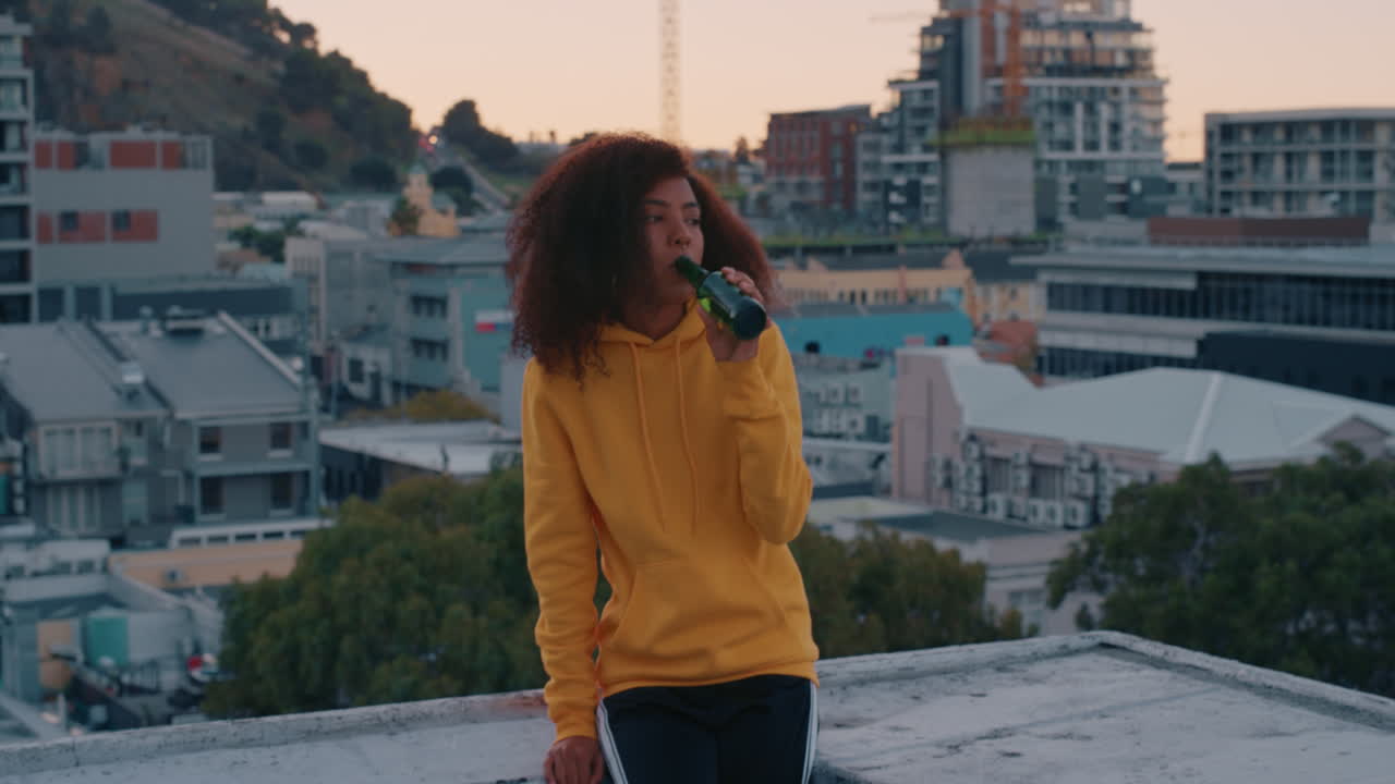 slow motion giovane donna afroamericana solitaria con stile afro che beve alcol che sembra infelice sul tetto al tramonto