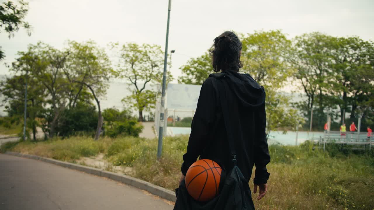 Rear view of a brunette man in a black jacket with a black backpack and a basketball goes to a basketball game on a street with a lot of trees
