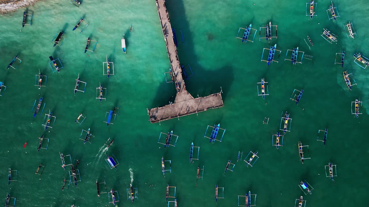 Overhead drone perspective of Bali fishing harbor capturing numerous small wooden boats anchored around a central jetty in shimmering turquoise waters under tropical daylight conditions