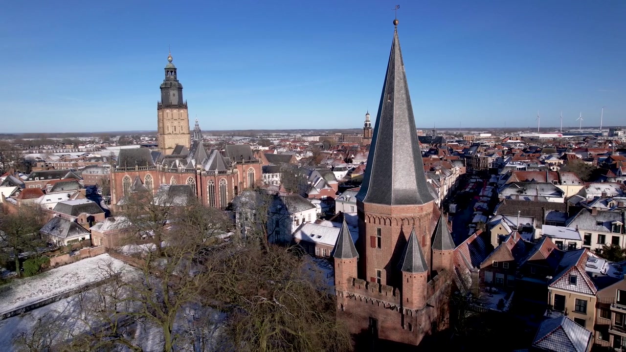 Movimento panoramico aereo cinematografico che mostra la storica città olandese con torre Zutphen Drogenapstoren, l'ingresso della città in primo piano e la cattedrale Walburgiskerk sullo sfondo, in un paesaggio invernale innevato in una giornata di sole.