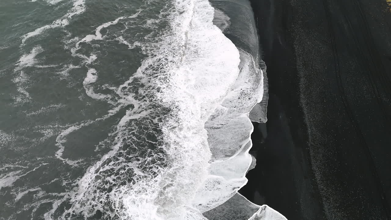 White foamy waves arrive to the beach with black sand in Iceland. Drone footage above the unusual shore of the ocean.