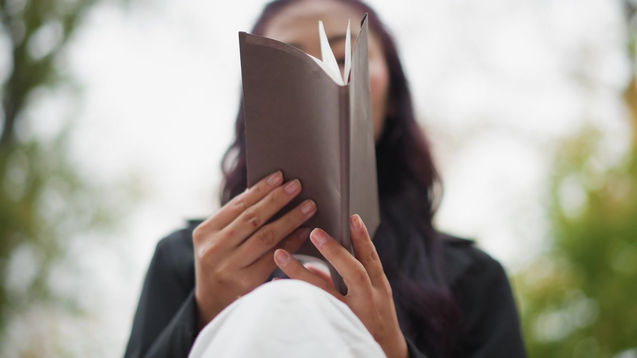 Primo piano di una giovane donna che tiene in mano e legge un libro aperto all'aperto. Le sue mani sono visibili mentre afferrano delicatamente la copertina marrone, mentre la donna si concentra sulle pagine. Lo sfondo è leggermente sfocato con elementi naturali.