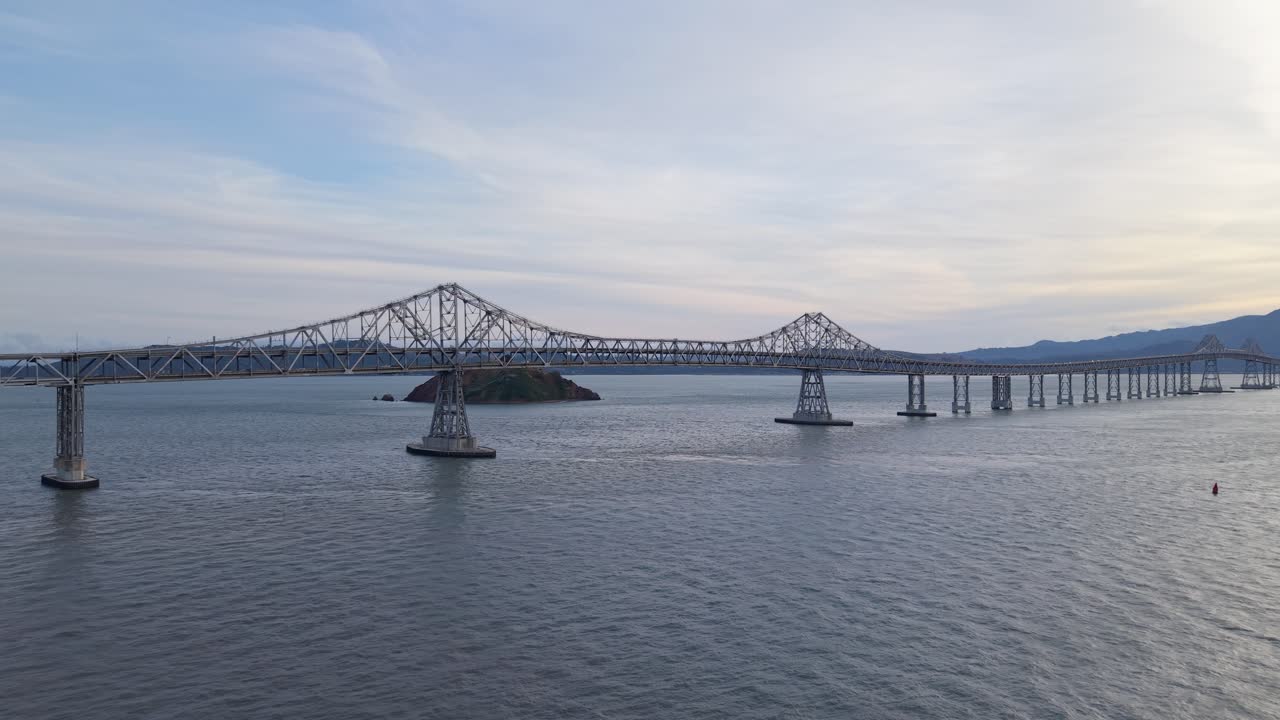 Drone view tracks across the bridge’s length, revealing steady traffic, expansive water, and layered clouds overhead. Filmed in 4K.