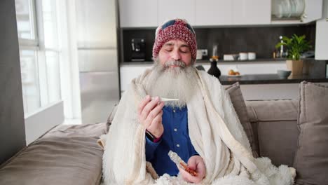 Portrait-of-an-elderly-man-in-a-red-cap-and-a-white-blanket-sits-and-checks-his-temperature-with-a-thermometer-and-also-holds-a-set-of-pills-in-his-hand-while-sitting-on-a-brown-sofa-in-a-modern-apartment