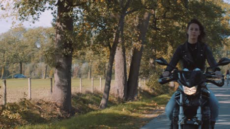 pan from forest fields, farmland to front view of beautiful european female biker wearing leather jacket on road with autumn leaf colored trees on sunny day