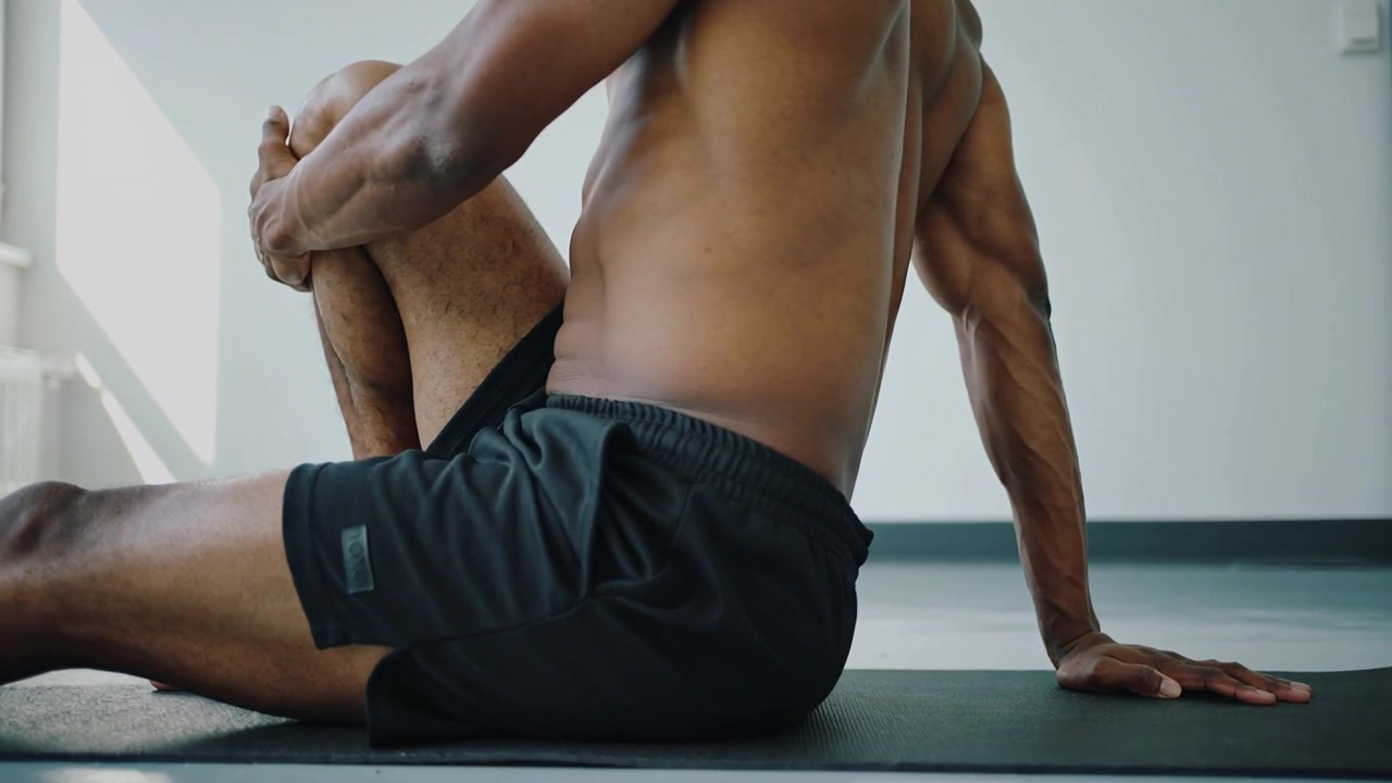 Acomodando-se no tapete, homem sem camisa de shorts pretos segurando o joelho esquerdo no peito, alongando-se no estúdio